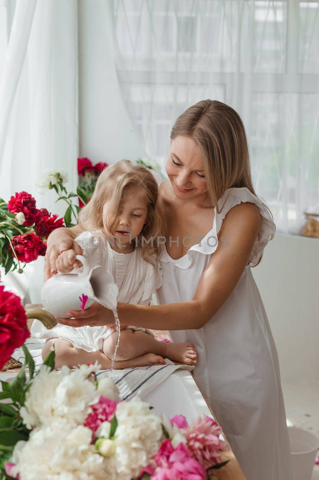 A little blonde girl with her mom on a kitchen countertop decorated with peonies. The concept of the relationship between mother and daughter. Spring atmosphere