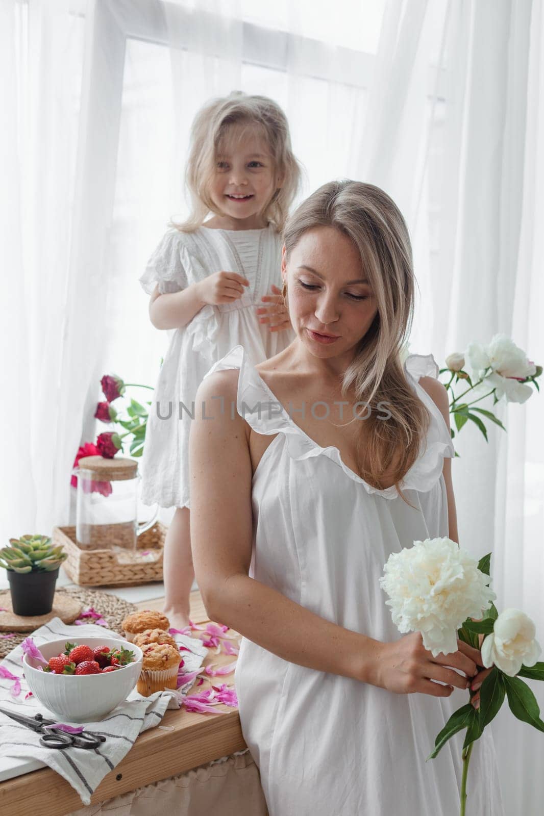 A little blonde girl with her mom on a kitchen countertop decorated with peonies. The concept of the relationship between mother and daughter. Spring atmosphere