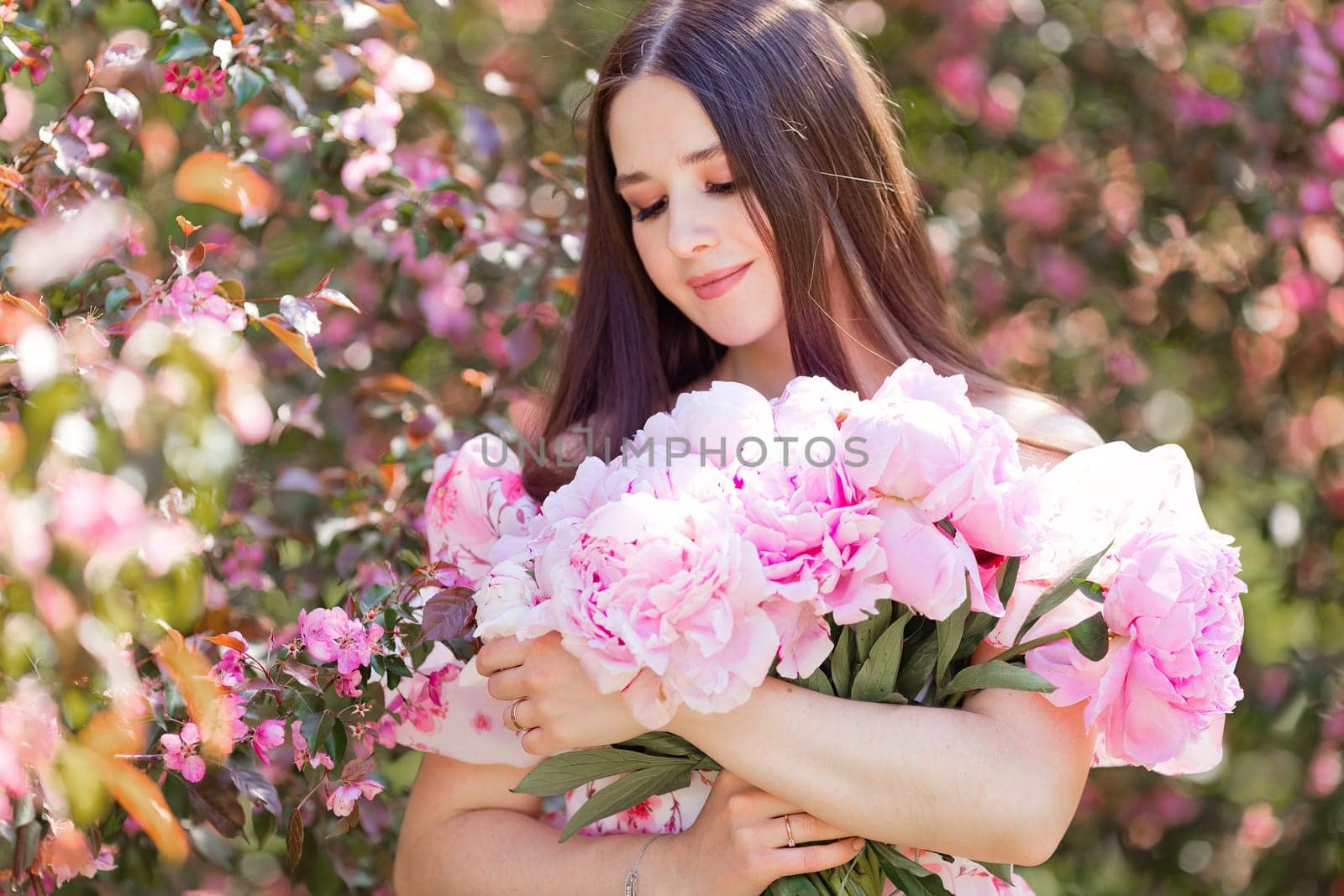A portrait of a smiling beautiful girl holding a large bouquet of pink peonies in her hands stands in a blooming garden on a sunny day. Copy space