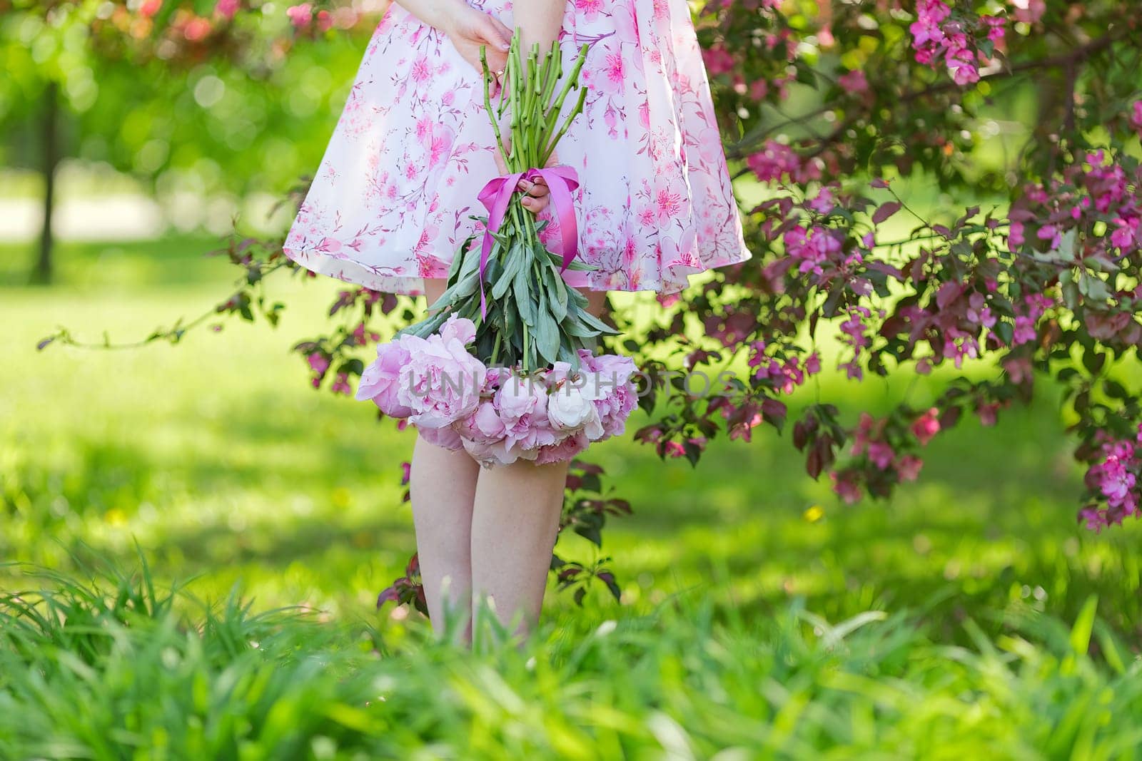 a girl in a pink dress, standing with a large bouquet of peonies on green grass, in a blooming garden. Back view. Copy space