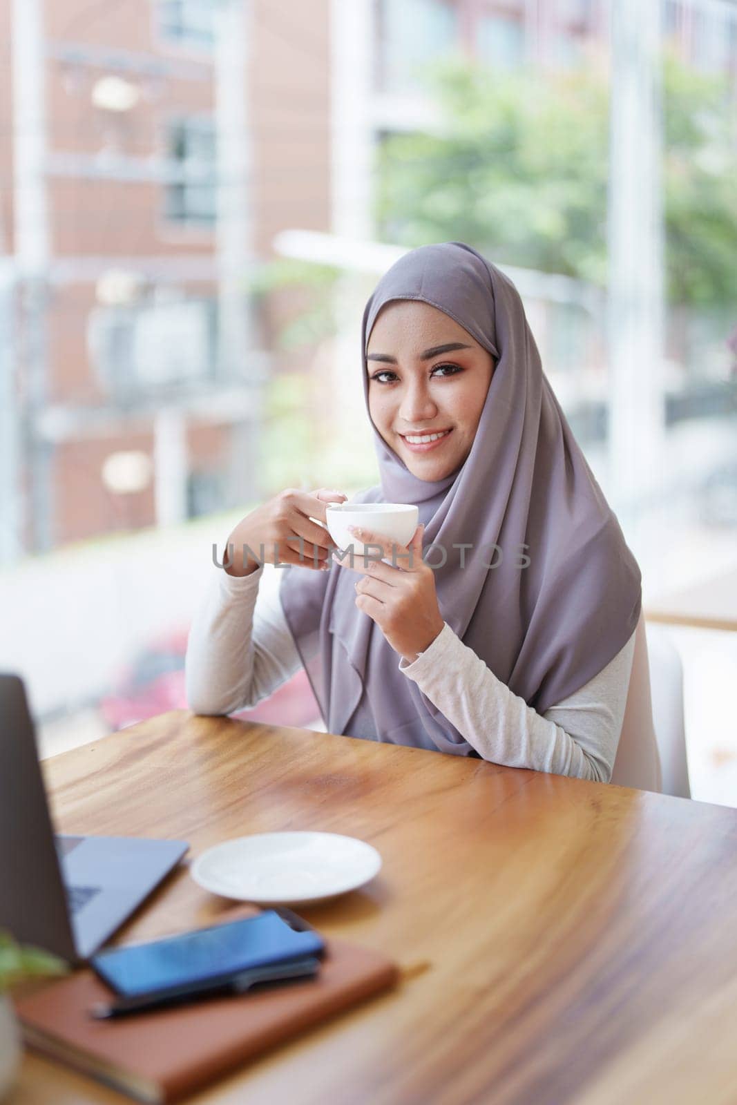 A beautiful Muslim woman with a smiling face in the morning drinking coffee and using a computer.