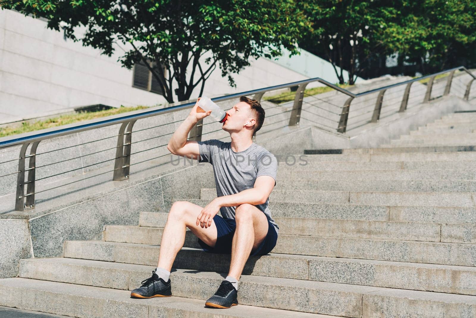 Tired Young man runner sitting on stairs and relaxing after sport training. Drinking water bottle while doing workout in summer city street