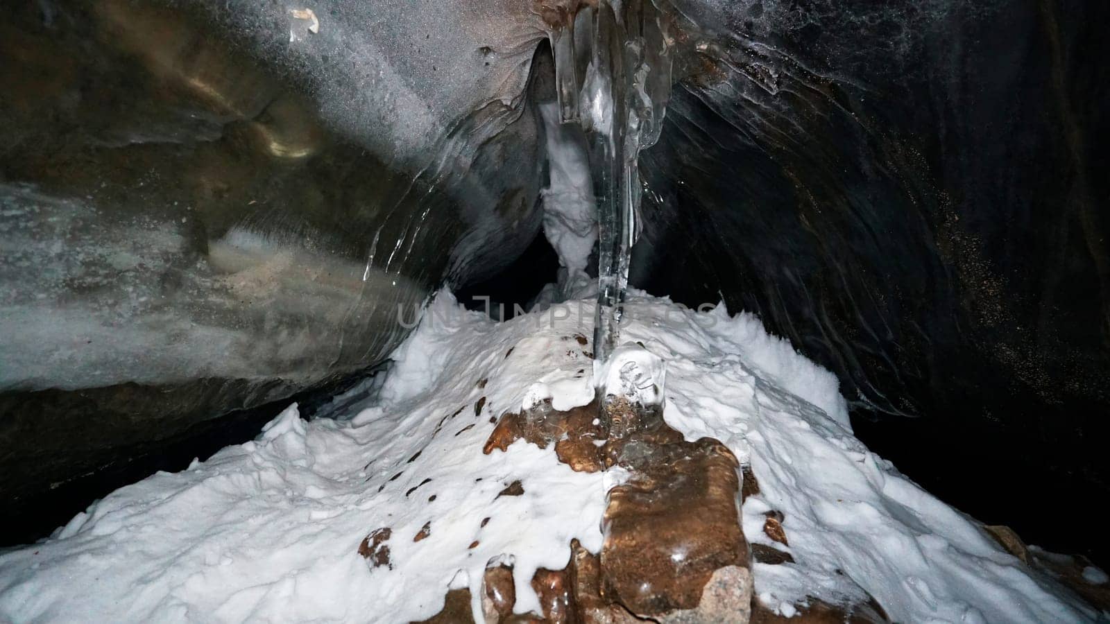 The black transparent color of ice in the cave. Small stones hang down in places. There are frozen air bubbles and large rocks in the ice. There is white snow. Icicles hang down. Small grains of snow