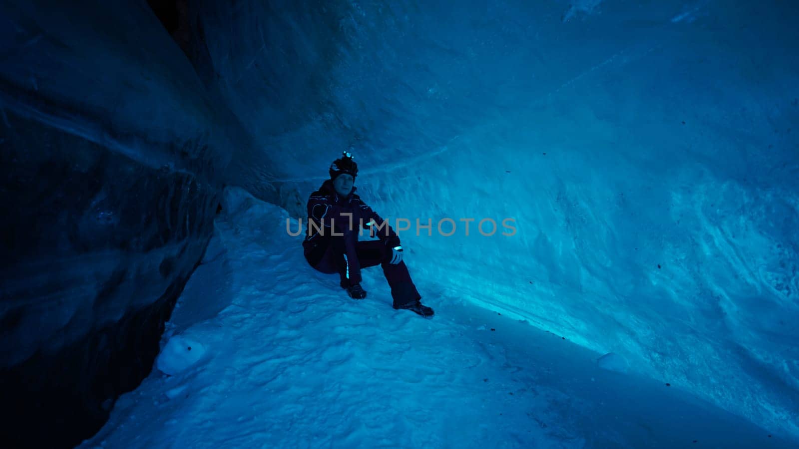 The climber made a halt inside an ice cave. The turquoise color of the ice gives a special atmosphere. The guy is looking for something in his backpack. There is snow on the icy floor. In mountains