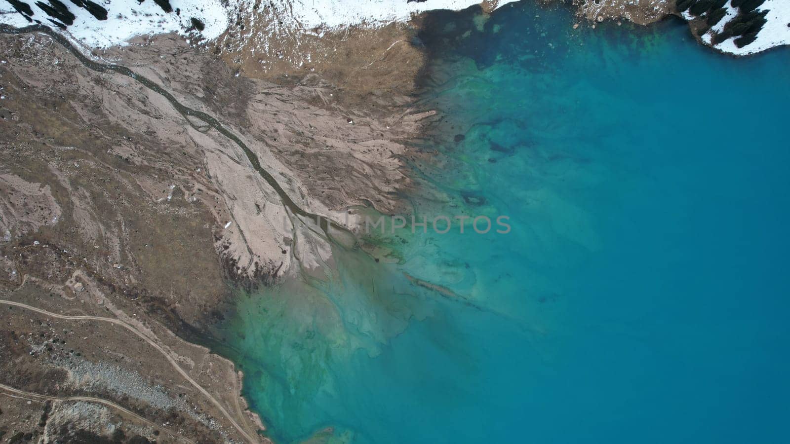 A lake in the mountains with turquoise blue water. Drone view of clear water, coniferous trees and snowy mountains. People walk along the shore, low bushes grow. Big Almaty lake. Kazakhstan