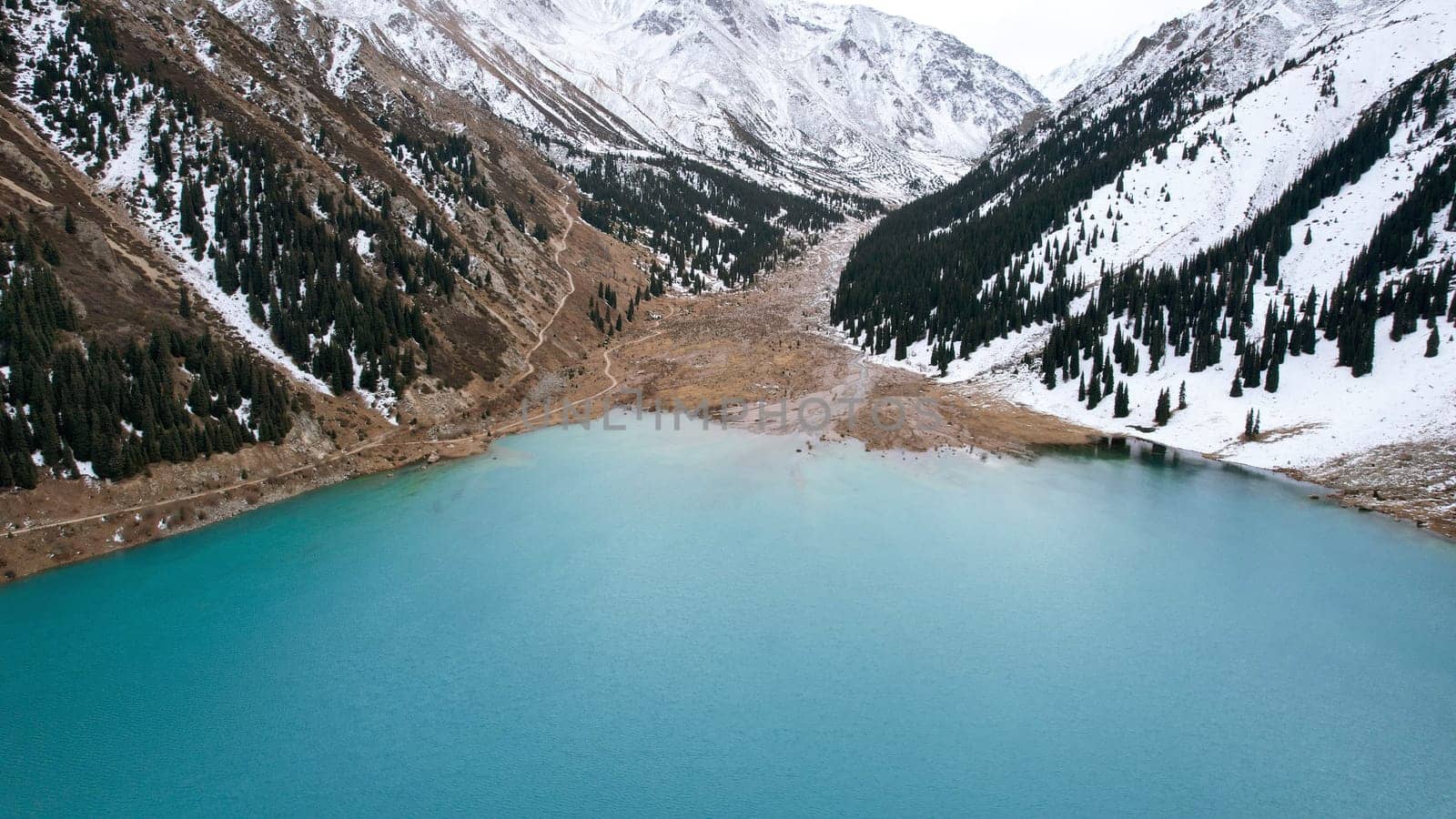 A lake in the mountains with turquoise blue water. Drone view of clear water, coniferous trees and snowy mountains. People walk along the shore, low bushes grow. Big Almaty lake. Kazakhstan