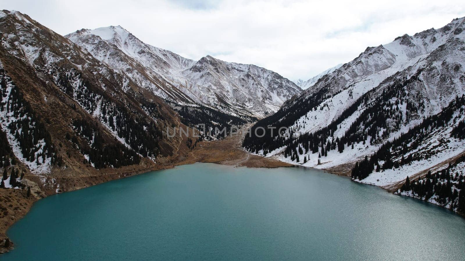 A lake in the mountains with turquoise blue water. Drone view of clear water, coniferous trees and snowy mountains. People walk along the shore, low bushes grow. Big Almaty lake. Kazakhstan