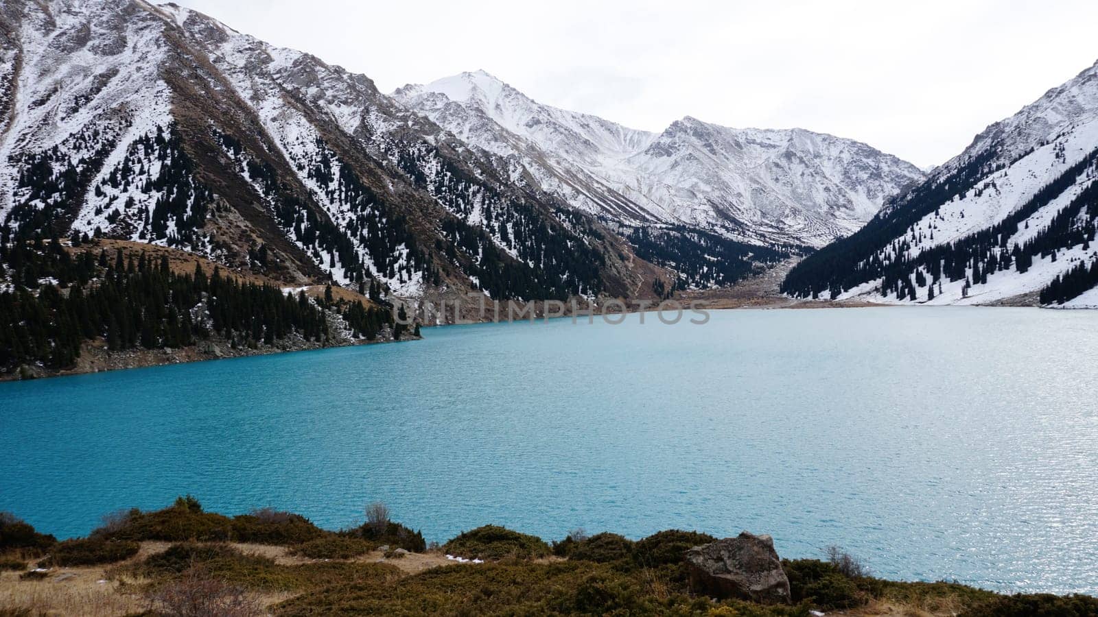 A mountain lake with blue water in winter. The sun's rays are reflected from the water. Green forest and grass grow on the hills. There are stones lying. The high peaks are covered with snow. Almaty