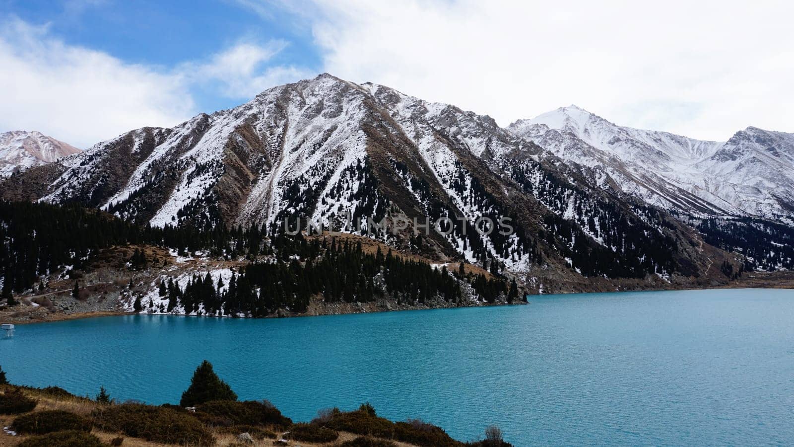 A mountain lake with blue water in winter. The sun's rays are reflected from the water. Green forest and grass grow on the hills. There are stones lying. The high peaks are covered with snow. Almaty