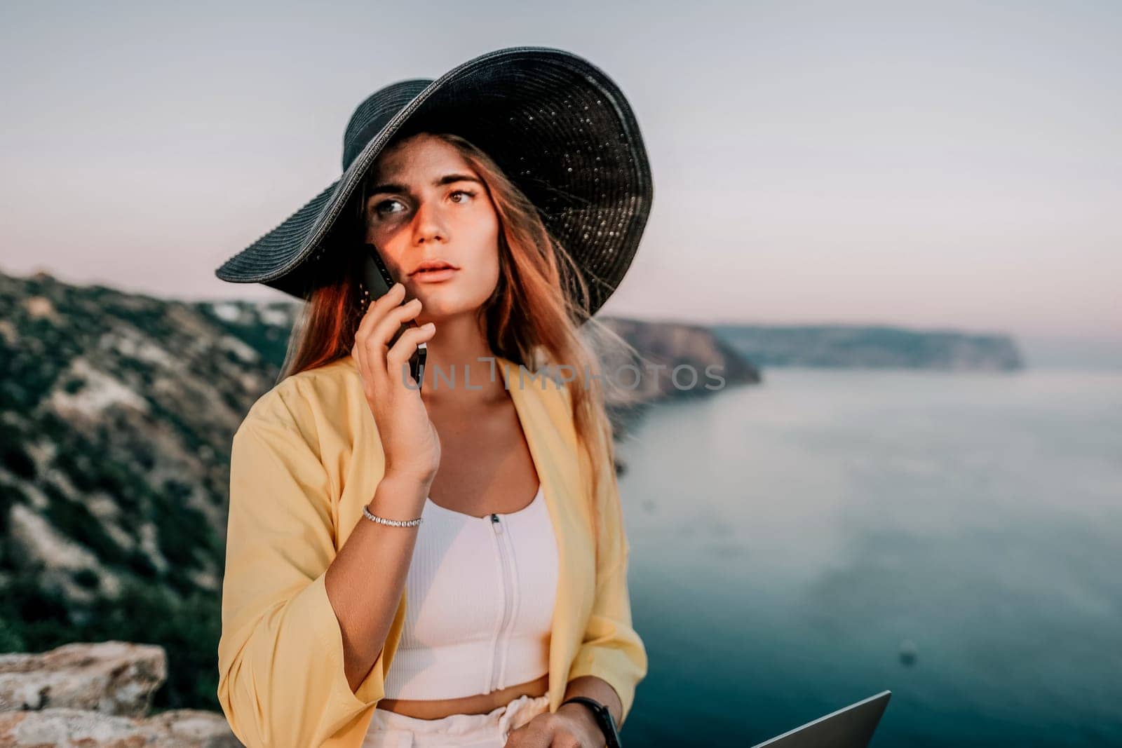 Successful business woman in yellow hat working on laptop by the sea. Pretty lady typing on computer at summer day outdoors. Freelance, travel and holidays concept.