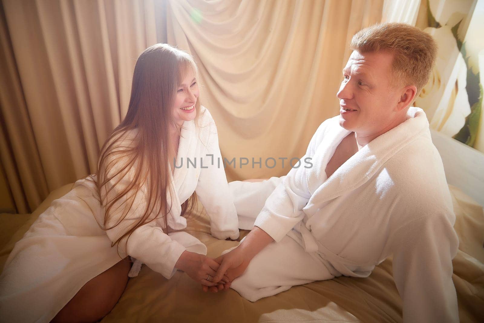 An adult couple of newlyweds in in white robes in a hotel room after the wedding. A guy and a girl, a man and a woman in beautiful room. The concept of hugs, love and care