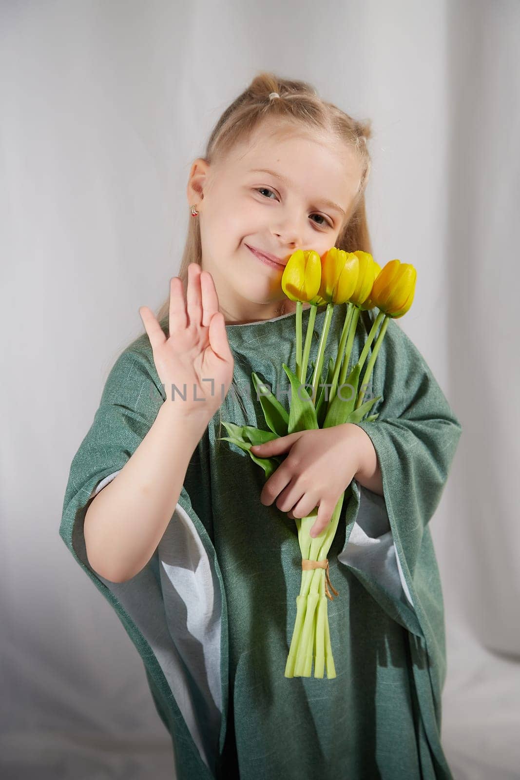 Portrait of a little blonde girl with a bouquet of spring yellow flowers on a light background. Child in green dress holding a bouquet of tulips in hands. Spring concept by keleny