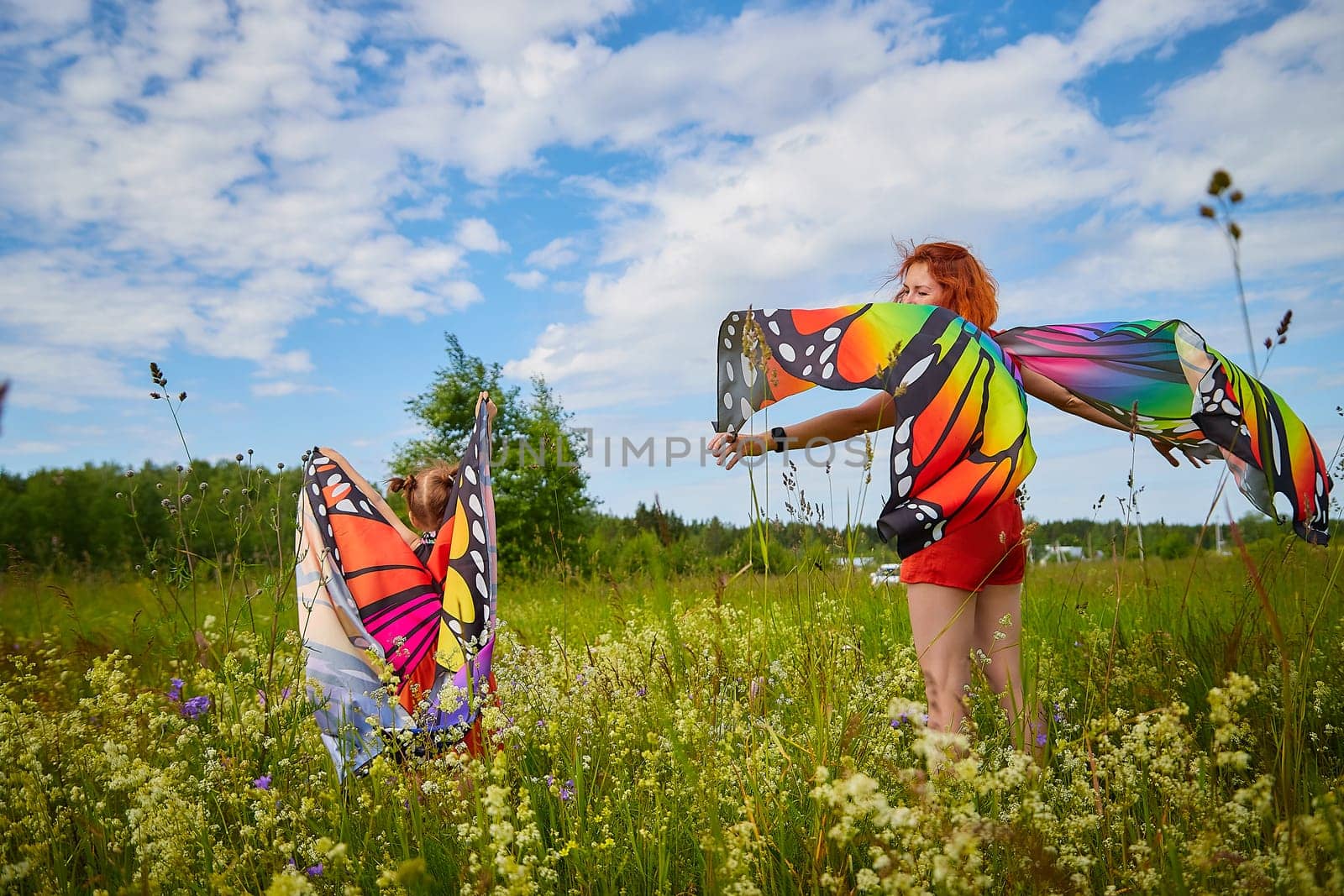 Happy female family with red haired mother and daughter with bright butterfly wings having fun on green and yellow meadow full of grass and flowers in sunny summer day. Concept family love