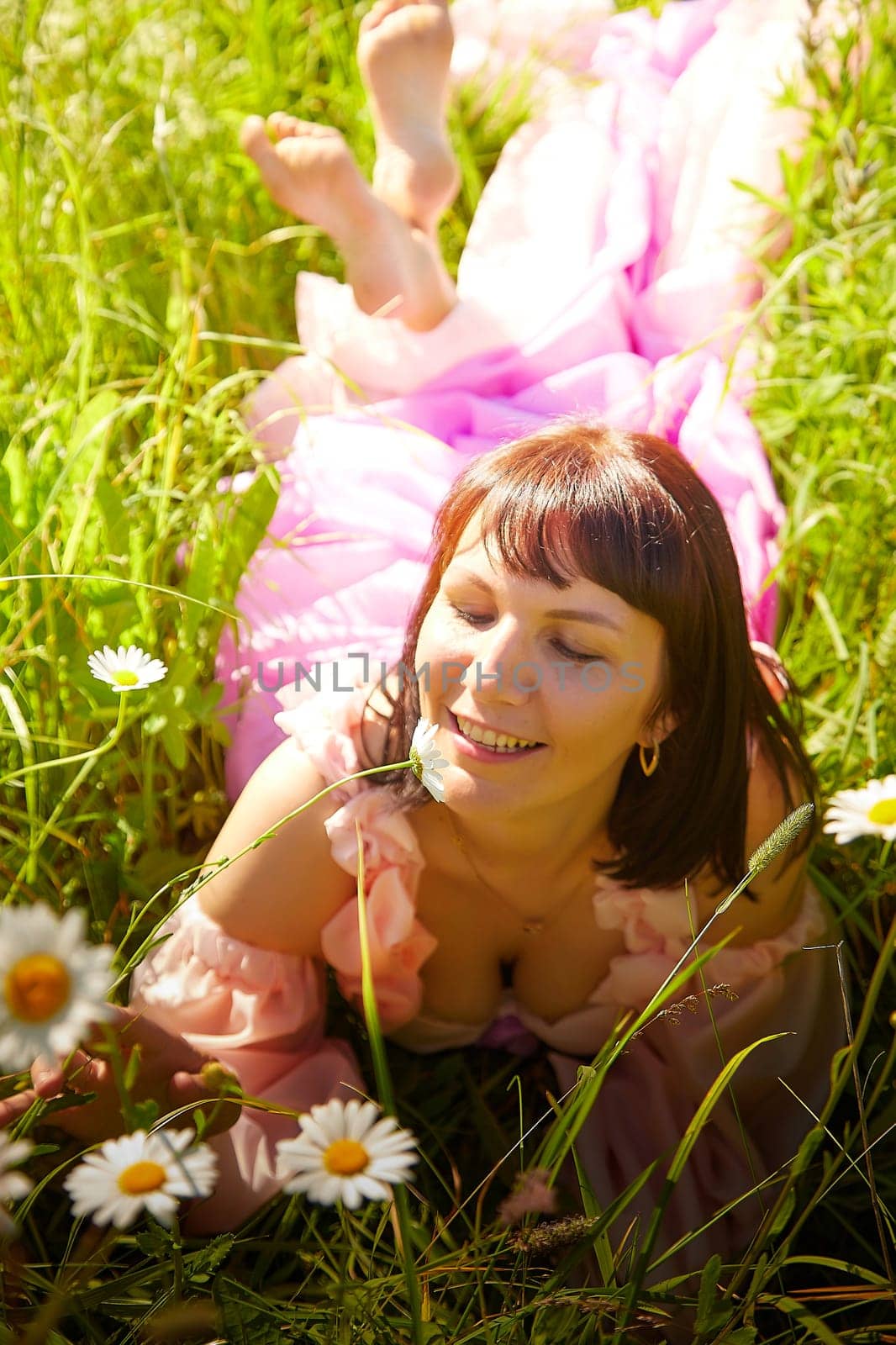 Beautiful girl in lush pink ball gown in green field during blooming of flowers and blue sky on background. Model posing on nature landscape as princess from fary tale