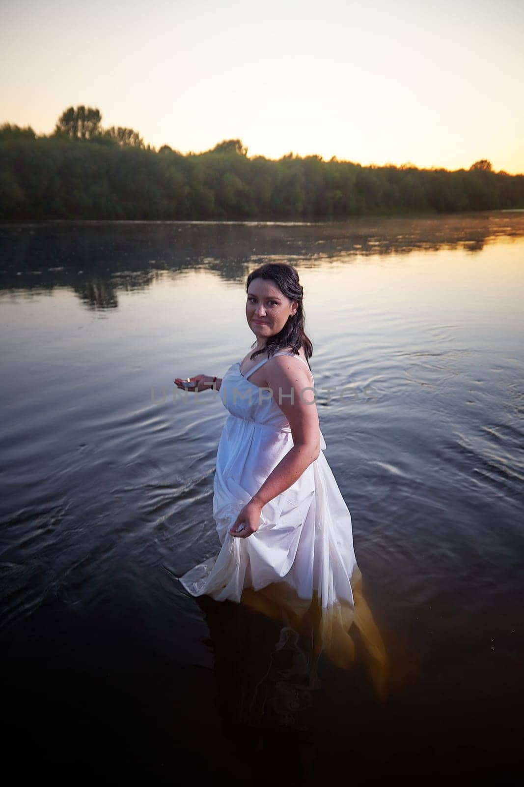 Slavic plump plump chubby girl in long white dress on the feast of Ivan Kupala with flowers and water in a river or lake on summer evening