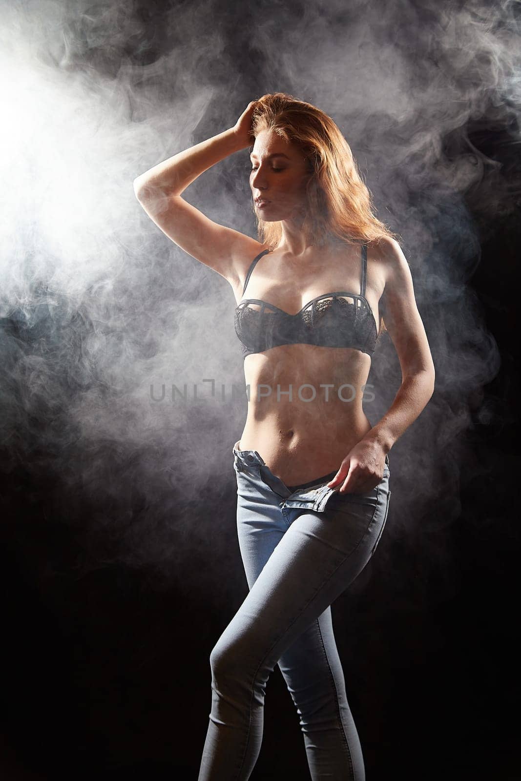 Luxury fitness girl with beautiful red hair stands in small black top and jeans taken off a pretty buttocks, alone in smoke in black background in photo studio by keleny