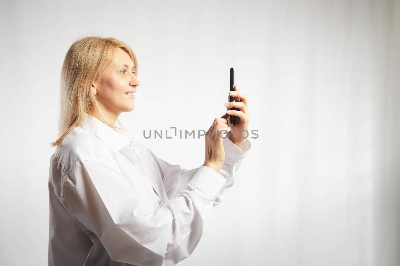 Portrait of a smiling casual blonde woman in shirt holding smartphone over white background. The girl is chatting and taking selfies. Business lady with a cell phone. Copy space by keleny