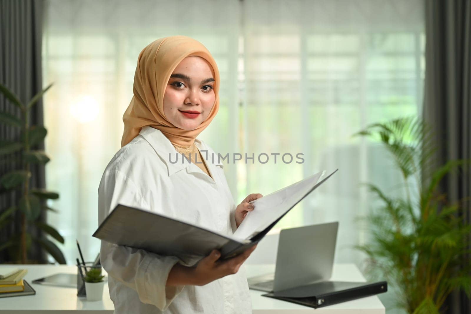 Image of professional muslim woman manger in hijab holding binder and smiling to camera.