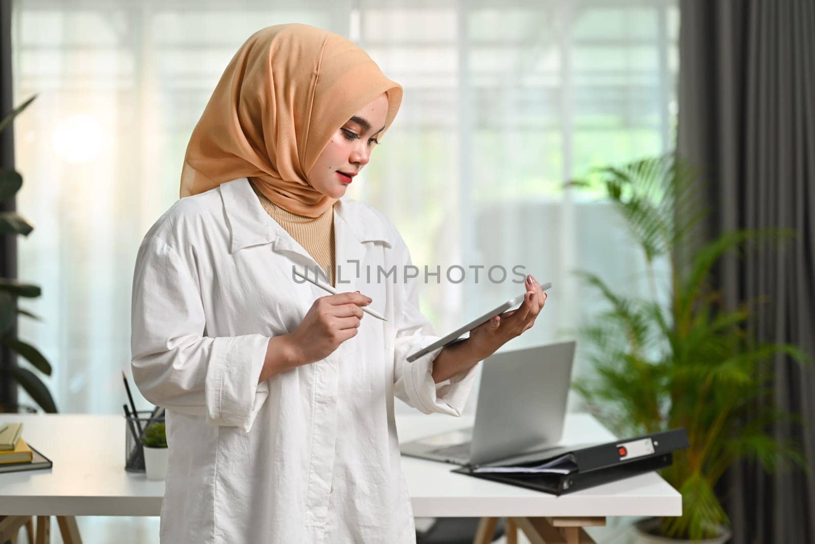 Muslim businesswoman wearing hijab using digital tablet while standing in an office boardroom.