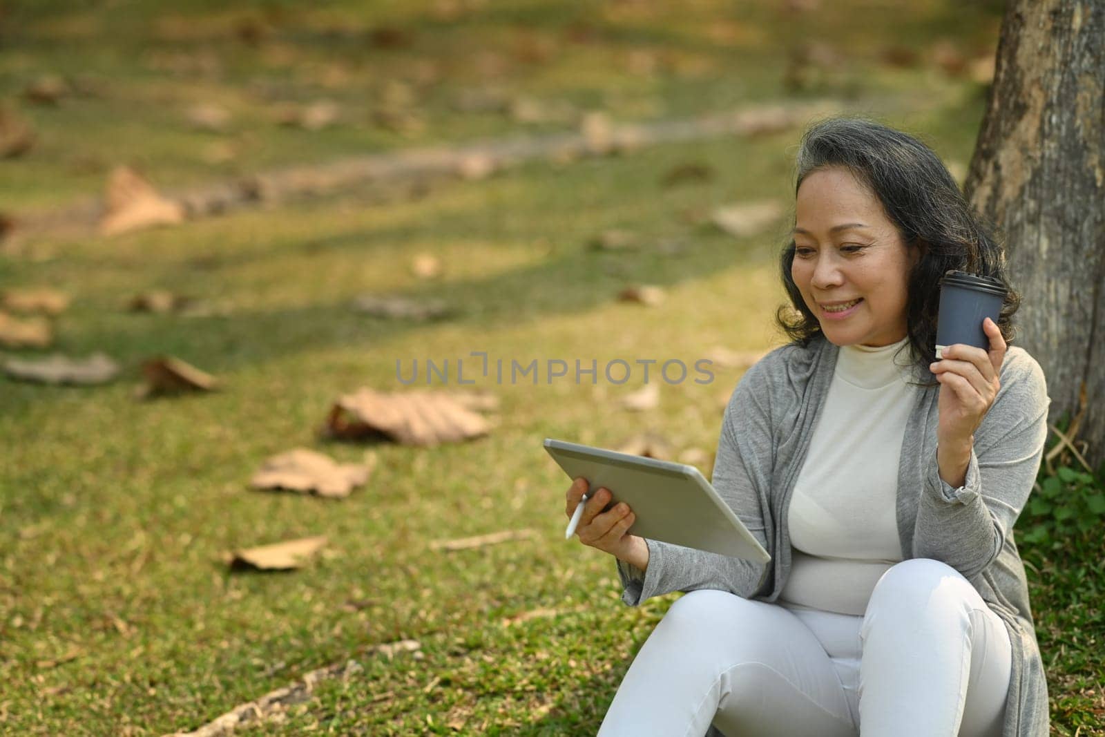 Positive middle aged woman holding paper cup of coffee using digital tablet under tree in the park at sunny beautiful day.