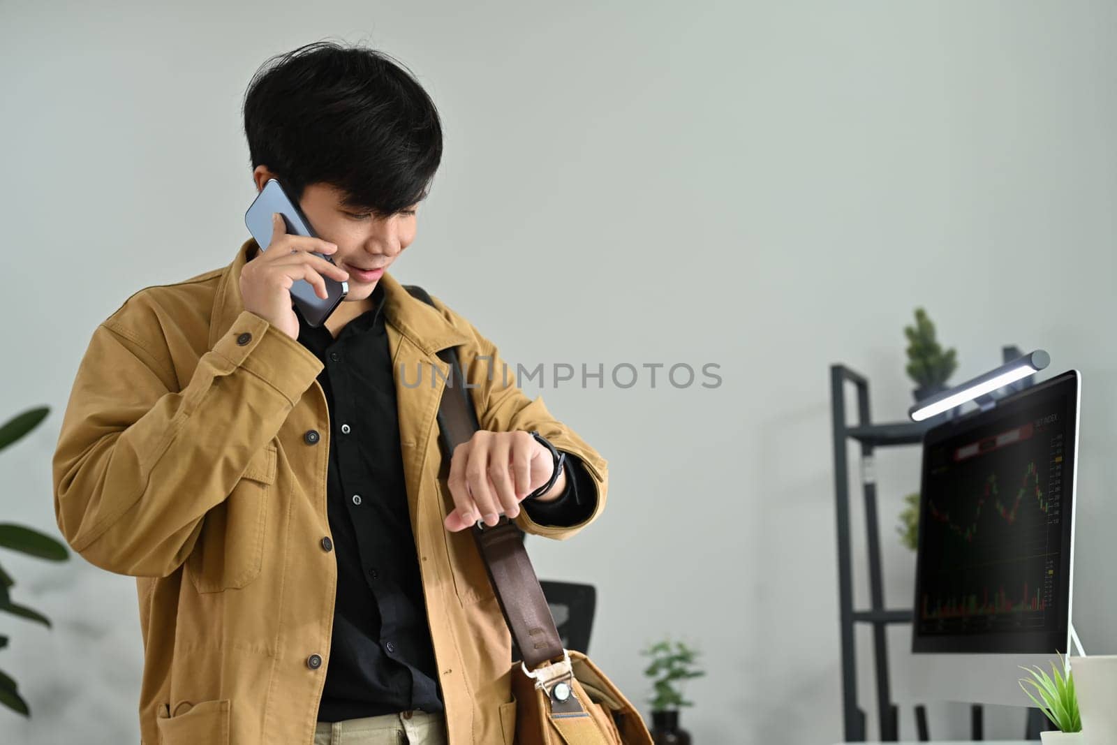 Image of young businessman or trader checking time on his watch and talking on mobile phone in modern office.