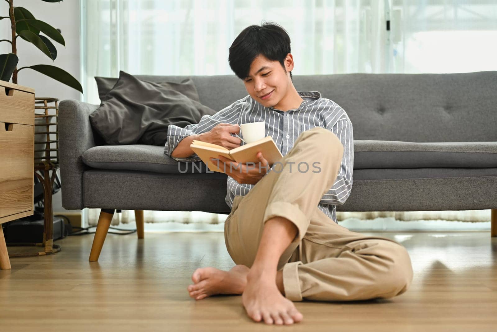 Full length of pleasant asian man sitting on floor and reading book, spending quality time at home.