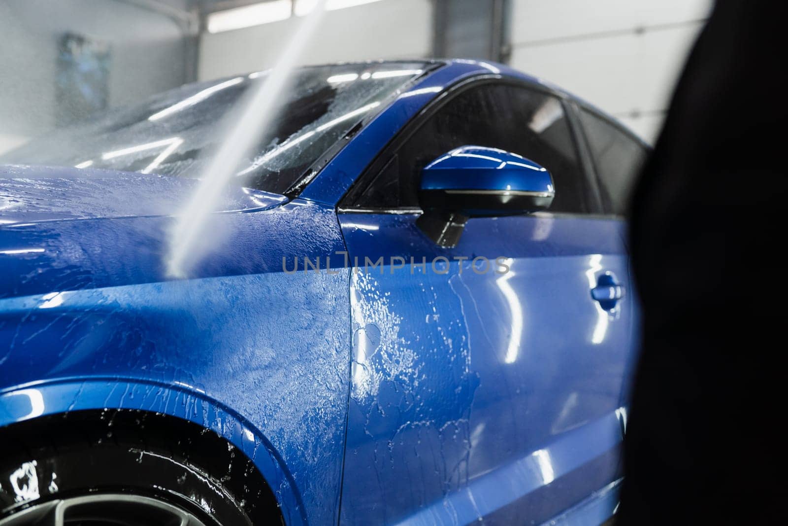 Car washer pouring water for full body car wash. Spraying water on car to wash off foam in the garage. Process of detailing washing in car service