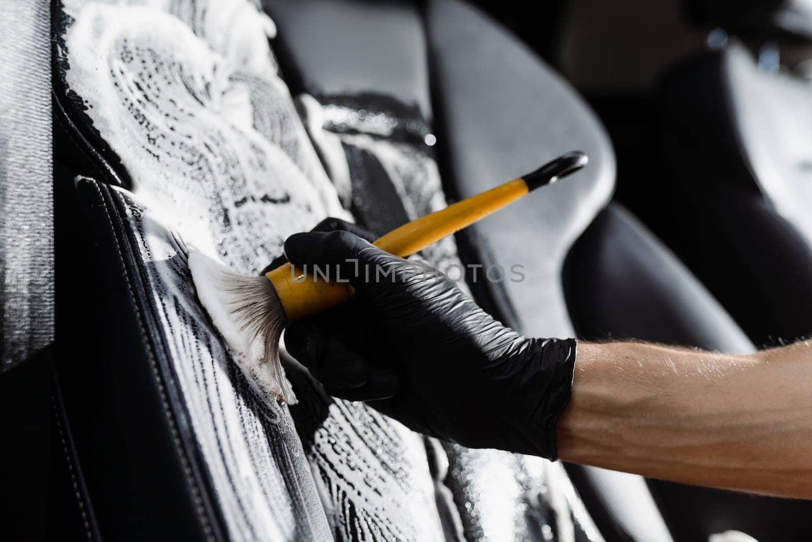 Foam cleaning black leather seat using brush. Worker in auto cleaning service clean car inside. Car interior detailing. by Rabizo