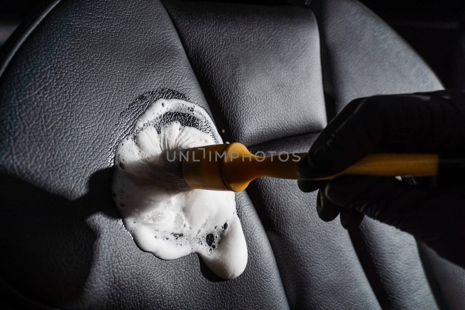 Process of foam and detergent cleaning leather seat using brush. Worker in auto cleaning service clean car inside. Car interior detailing. by Rabizo
