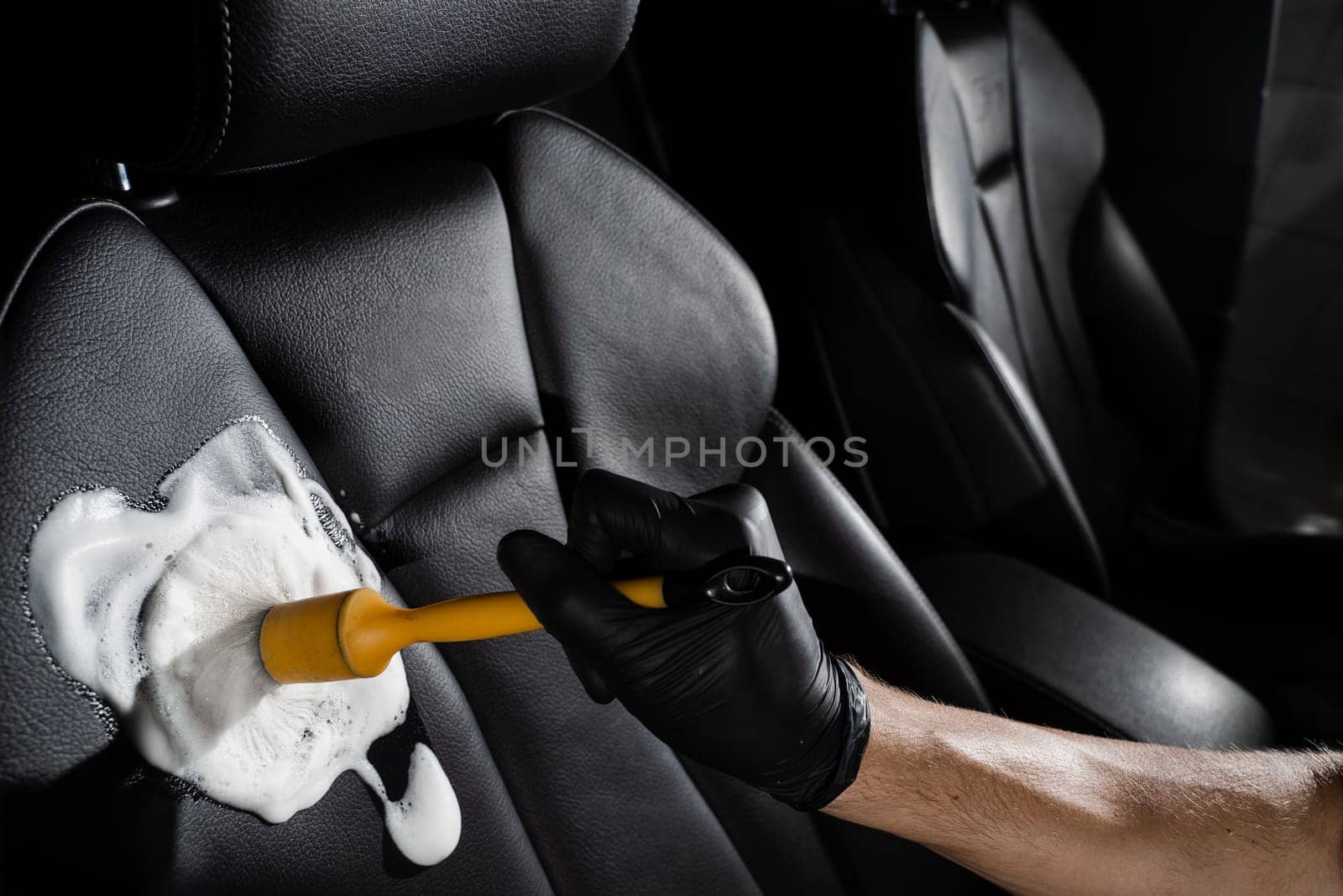 Process of foam and detergent cleaning leather seat using brush. Worker in auto cleaning service clean car inside. Car interior detailing. by Rabizo