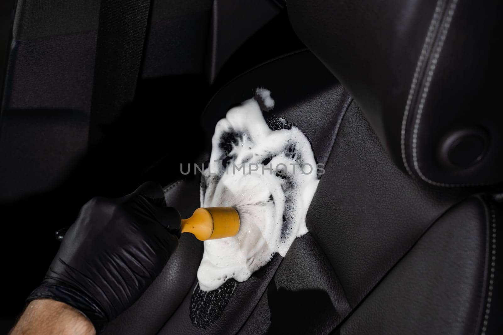Process of foam and detergent cleaning leather seat using brush. Worker in auto cleaning service clean car inside. Car interior detailing. by Rabizo