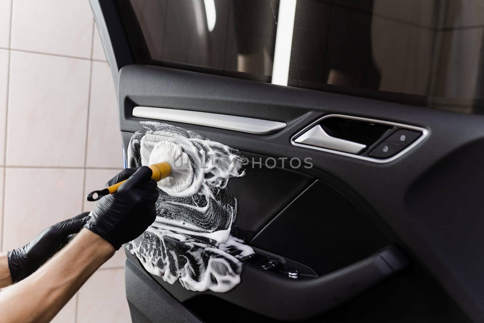 Process of foam and detergent cleaning with brush inside handle of car door. Car detailing service. Worker in cleaning service washing leather interior of clients auto. by Rabizo