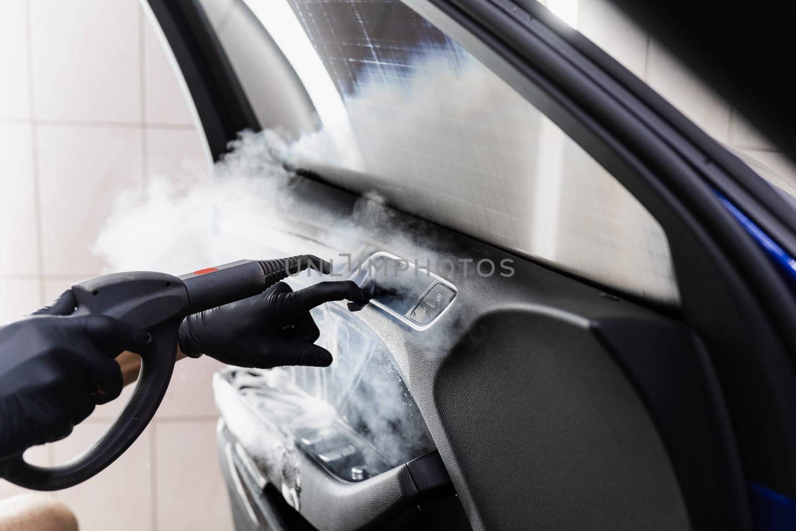 Steam vaping cleaning inside handle of car door after foam washing with brush. Worker in car detailing service washing leather interior of clients auto using steam generator. by Rabizo