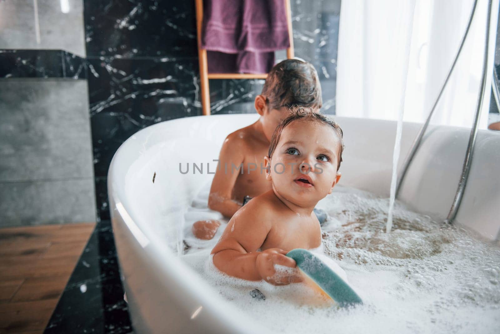 Two kids having fun and washing themselves in the bath at home. Helping each other.