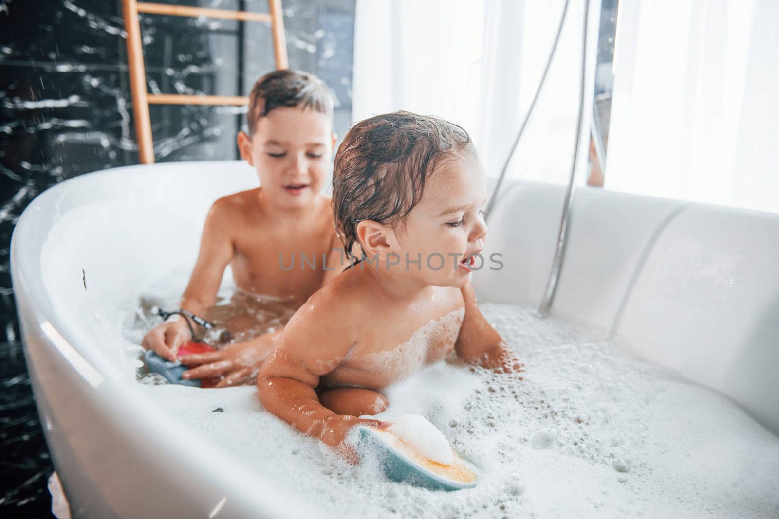 Two kids having fun and washing themselves in the bath at home. Helping each other.