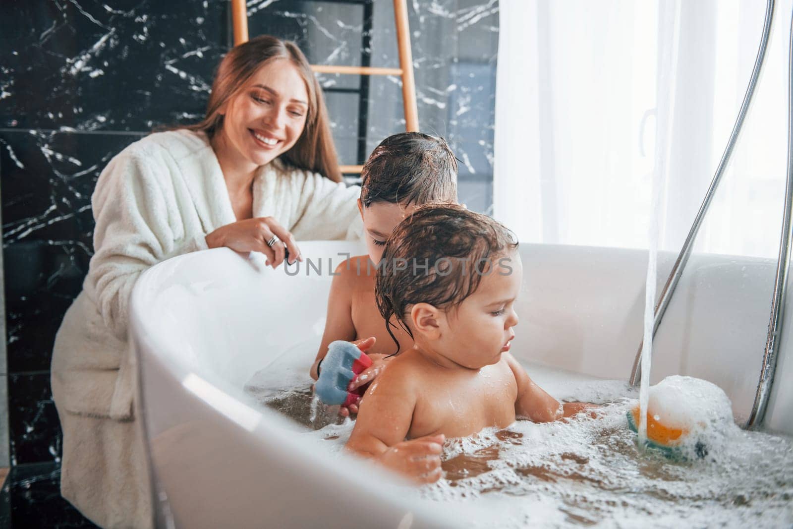 Young mother helps her son and daughter. Two kids washing in the bath.