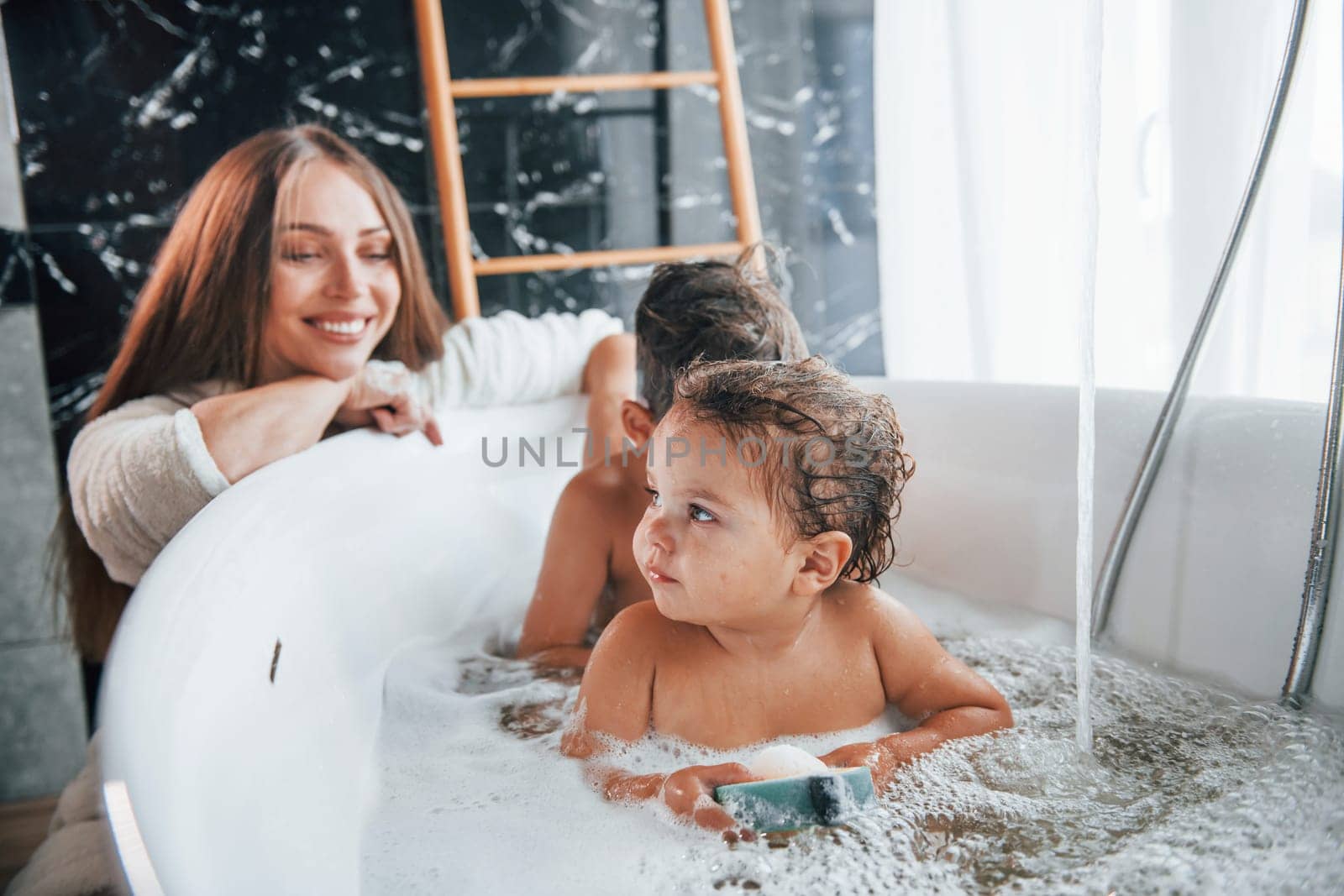 Young mother helps her son and daughter. Two kids washing in the bath.