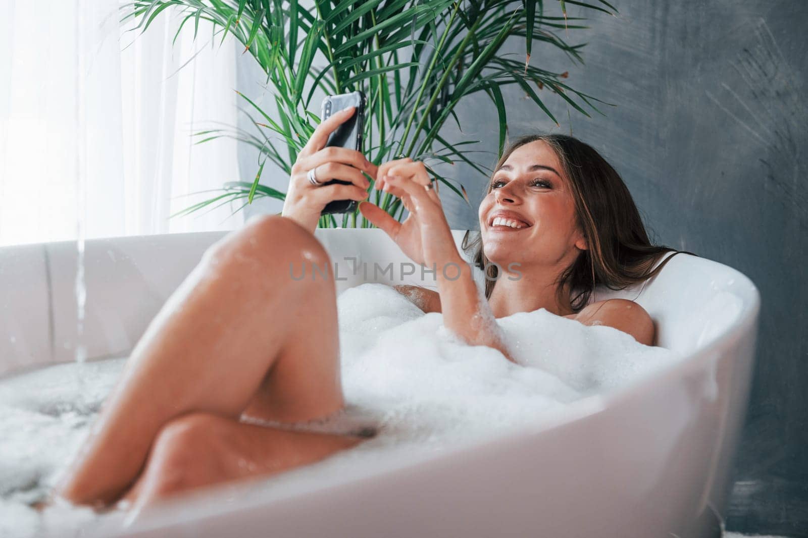 Uses phone. Beautiful young woman have free time at home in the bath.