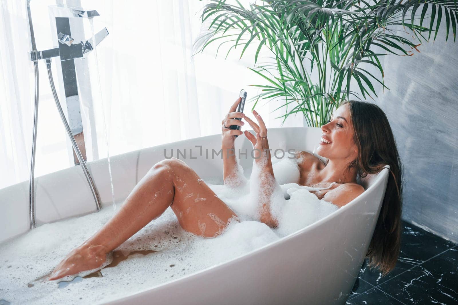 Uses phone. Beautiful young woman have free time at home in the bath.