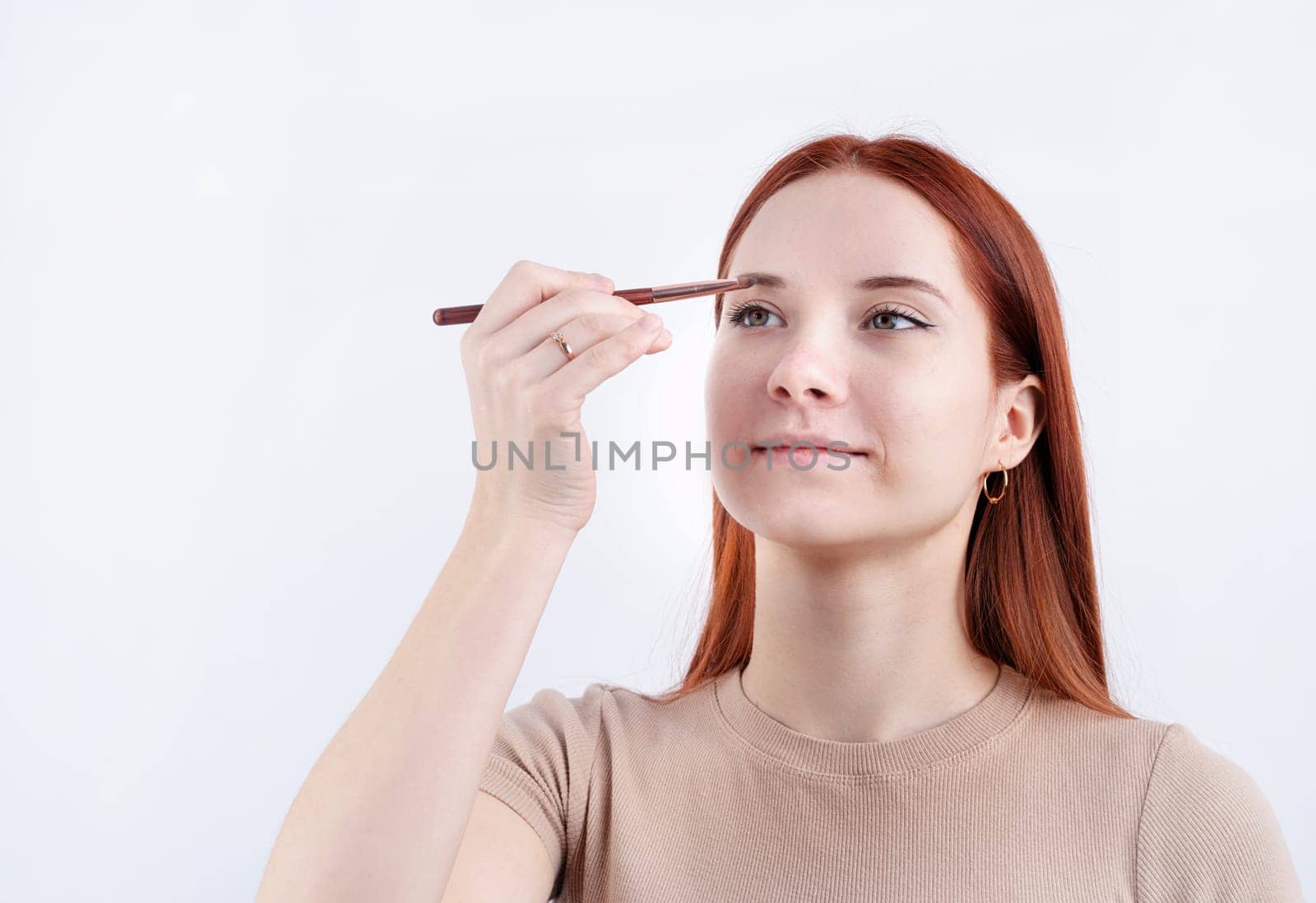 Joyful redhead young woman using makeup brush making up isolated on white background by Desperada