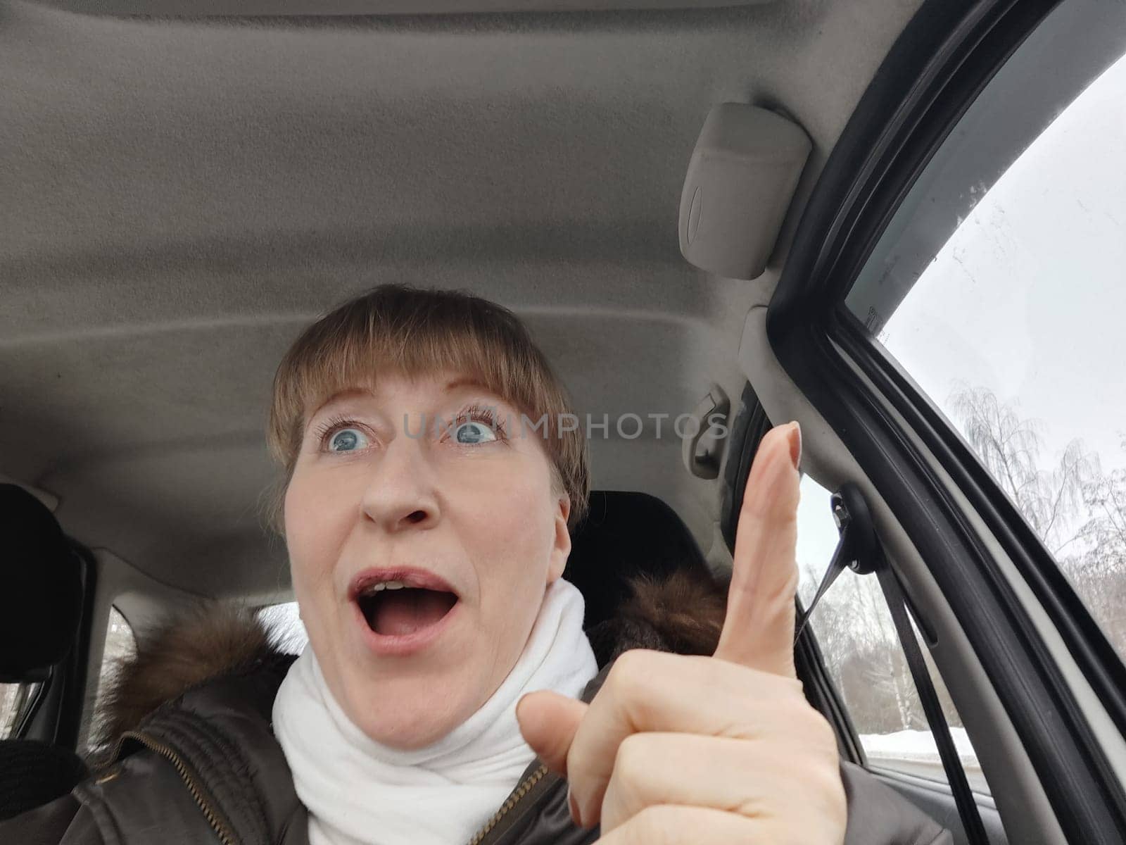 Portrait of adult cute funny blonde woman in warm clothes taking a selfie inside the car and driving it. The concept of pleasant trip in spring, autumn, winter