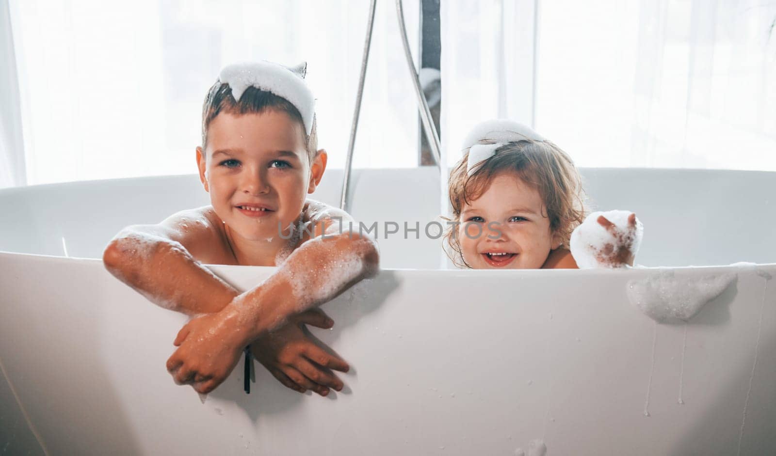 Two kids having fun and washing themselves in the bath at home. Posing for a camera.