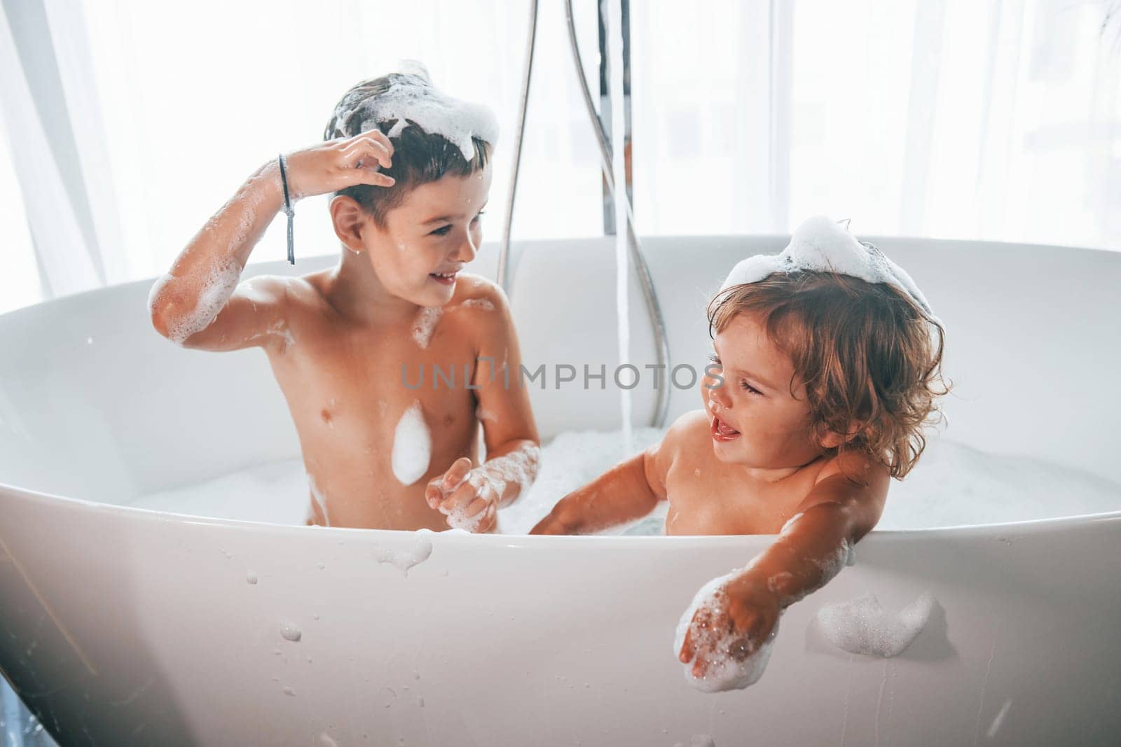 Two kids having fun and washing themselves in the bath at home.