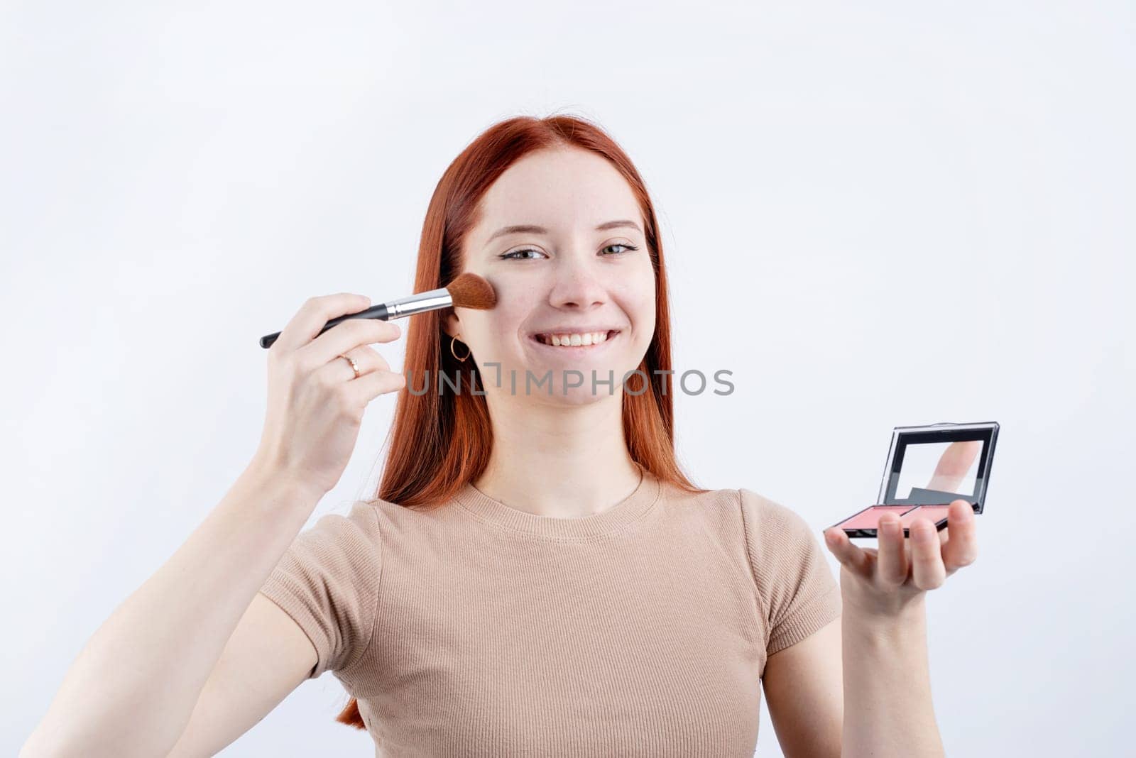 Joyful redhead young woman using makeup brush and blusher isolated on white background by Desperada