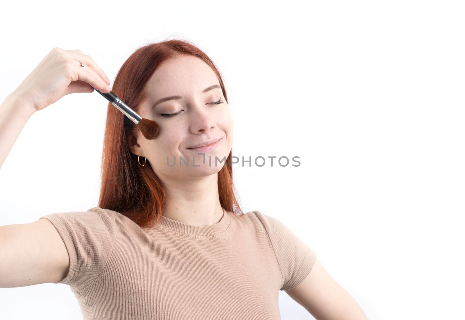 Joyful redhead young woman using makeup brush making up isolated on white background, copy space, natural beauty