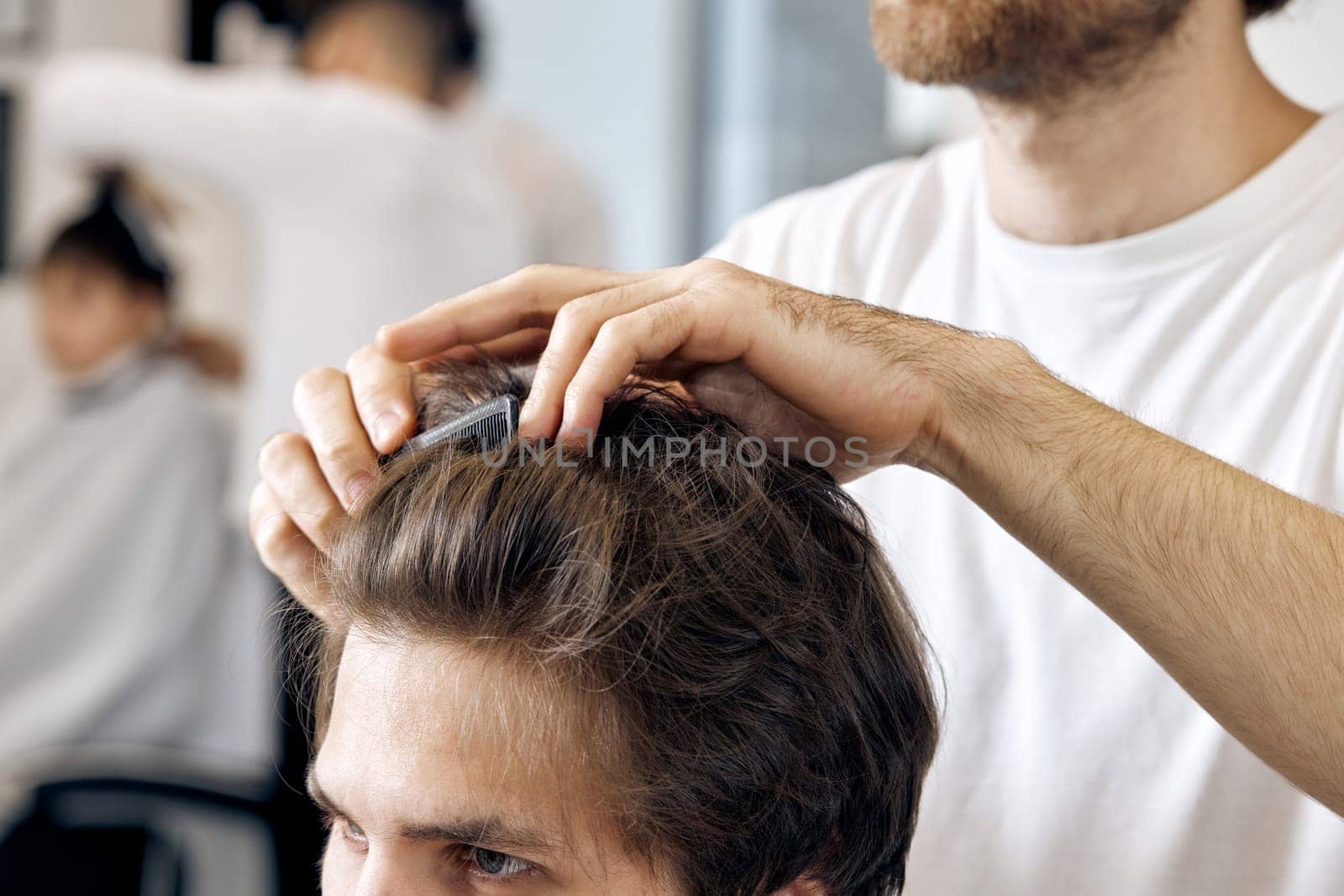 close-up, professional male hairstylist combing young customer's hair in barbershop