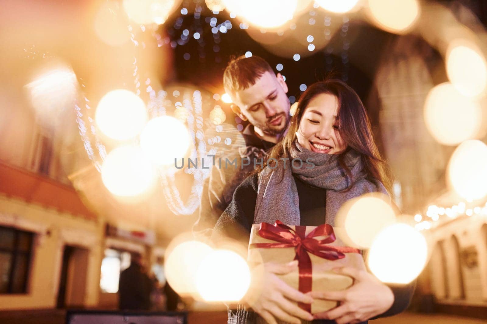 Holds gift box. Happy multiracial couple together outdoors in the city celebrating New year.