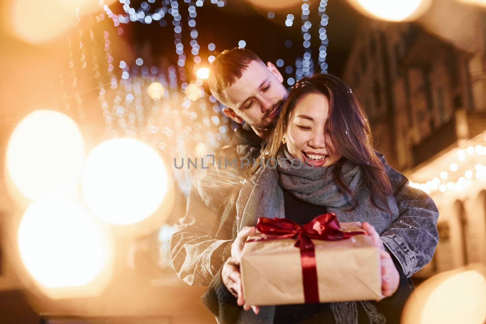 Holds gift box. Happy multiracial couple together outdoors in the city celebrating New year.