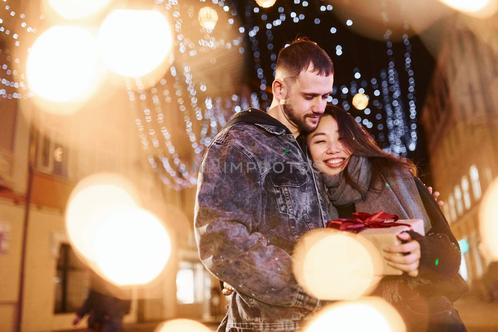 Holds gift box. Happy multiracial couple together outdoors in the city celebrating New year.