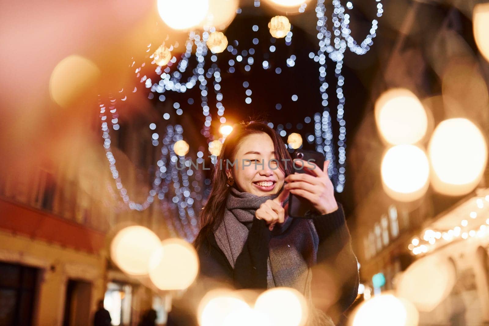 Uses phone. Cute and happy asian young woman outdoors in the city celebrates New year.